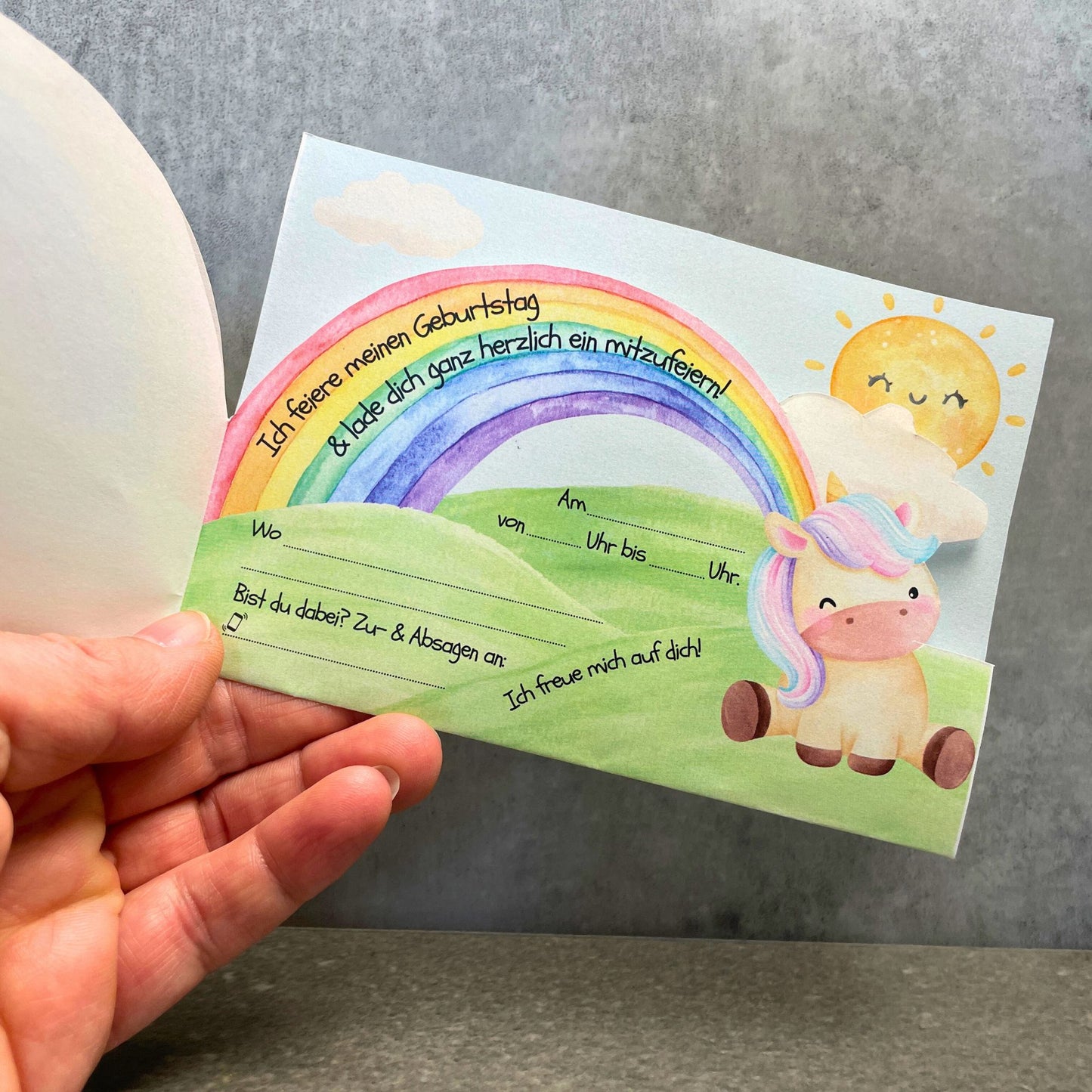 Einhorn Einladung Innenteil mit Einladungstext zum Ausfüllen. Grüne Hügellandschaft mit sitzendem Einhorn, buntem Regenbogen und freundlicher Sonne.